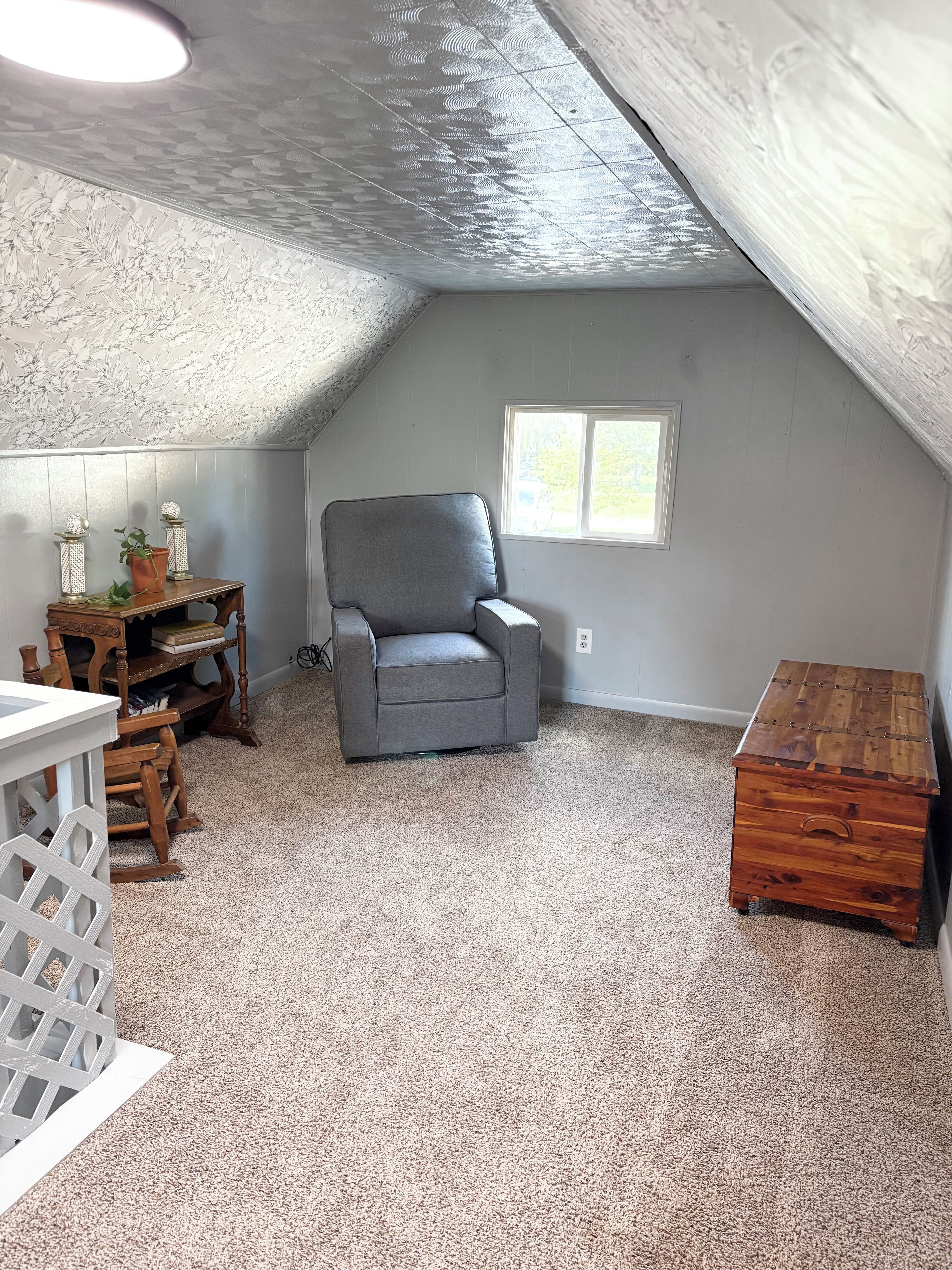 A room with a sloped ceiling, a grey upholstered chair, a wooden chest of drawers, and a small window.