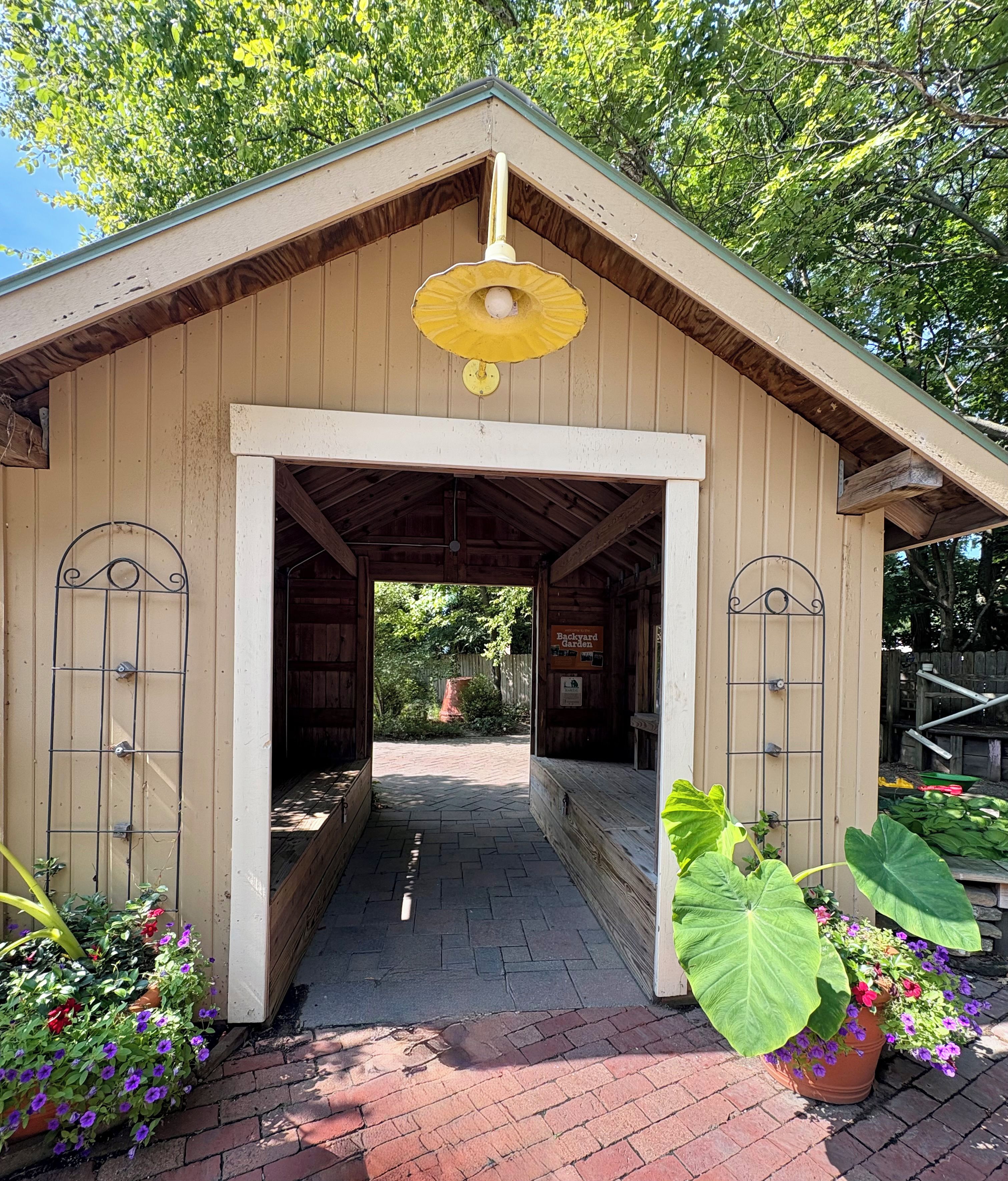 A wooden structure with a covered entrance, a sign indicating a 'Backyard Garden', and potted plants with flowers.