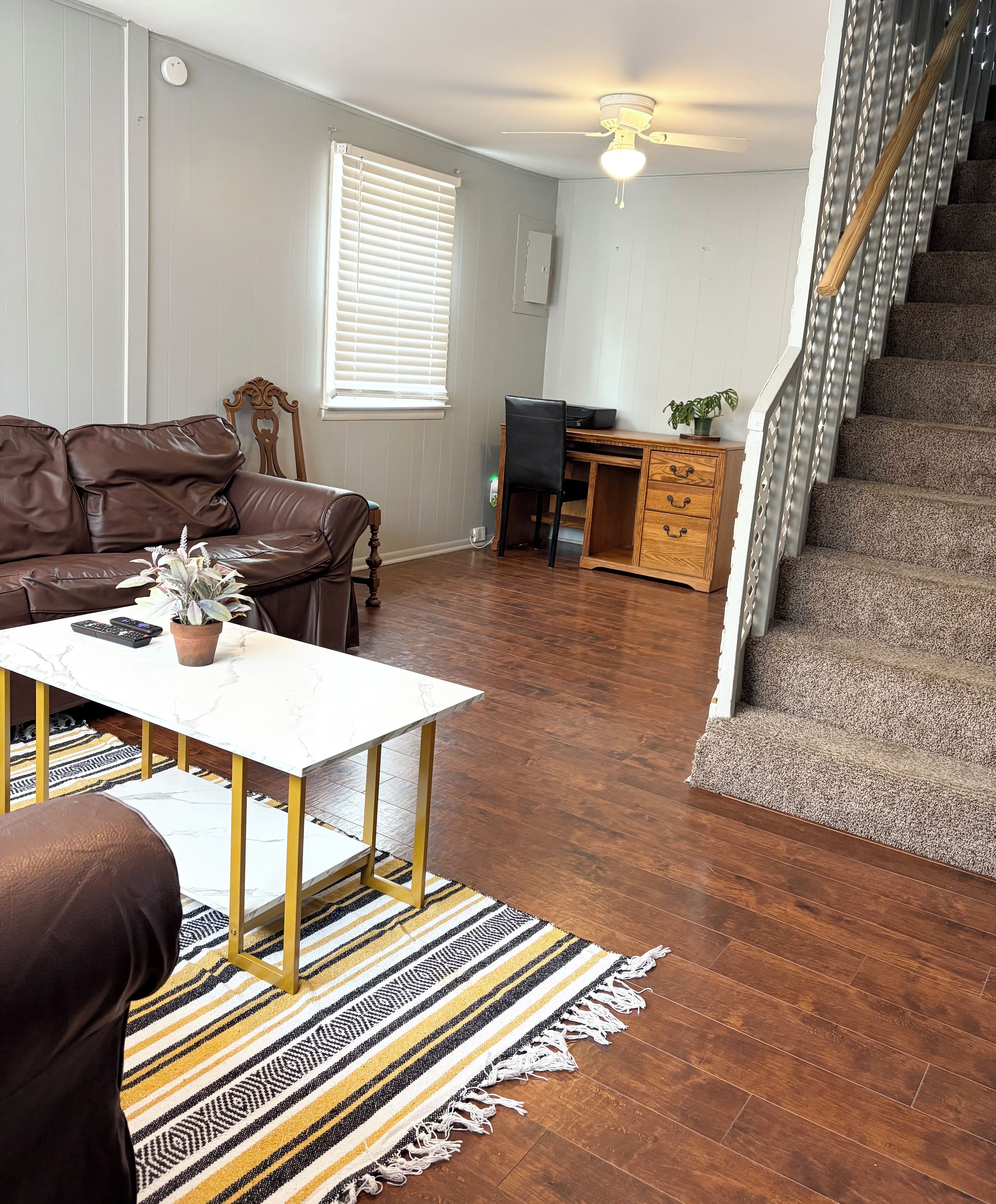 A living room with a brown leather sofa, a white coffee table, a striped rug, a wooden desk, and a staircase.