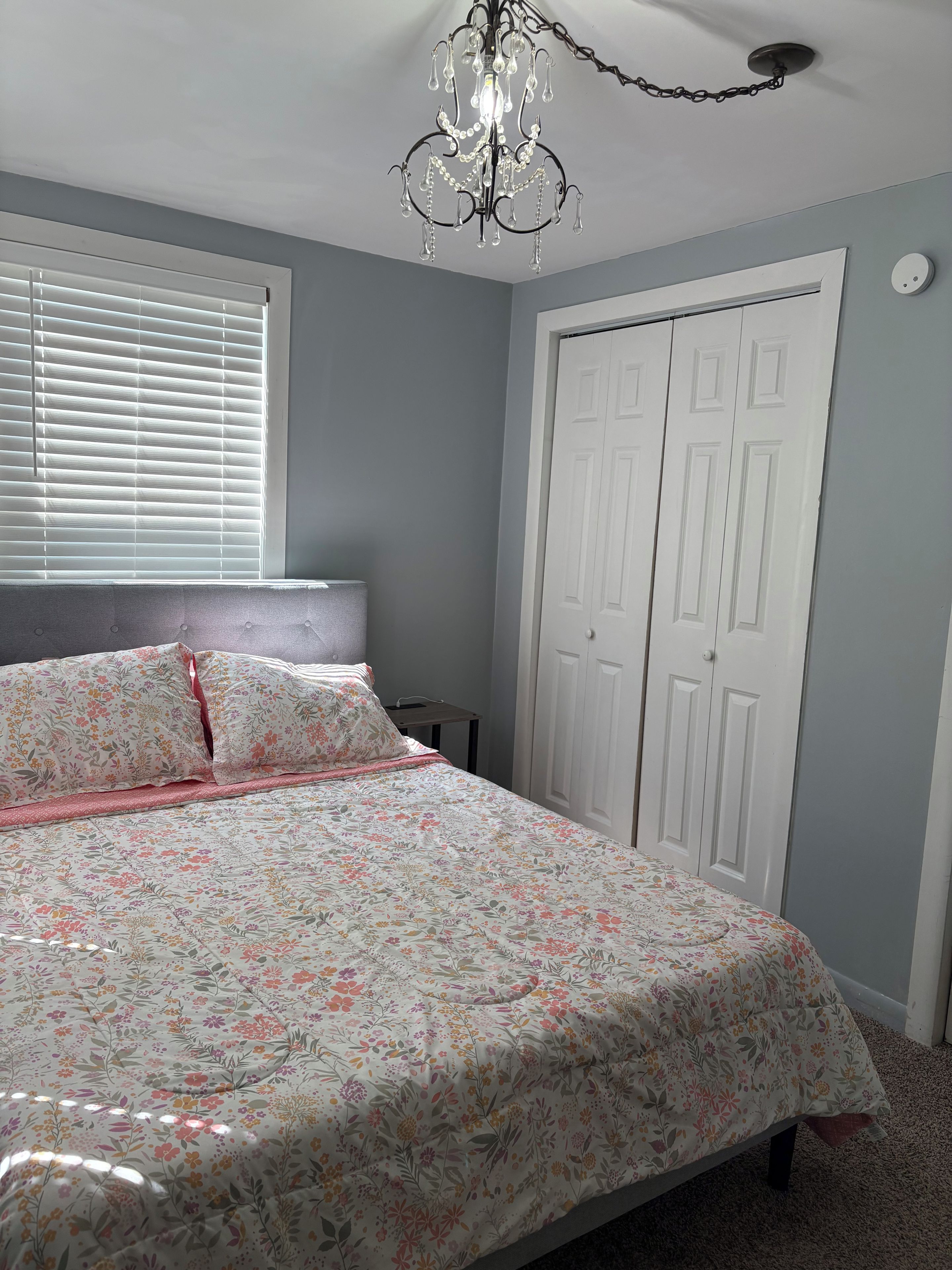 A bedroom with a bed, a window with blinds, a white wardrobe, and a chandelier.