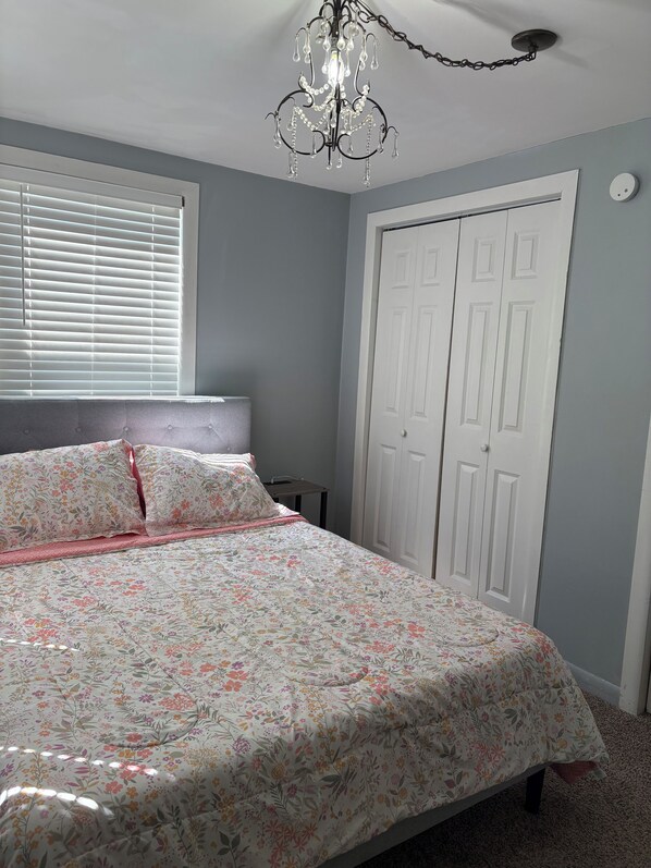 A bedroom with a bed, a window with blinds, a white wardrobe, and a chandelier.