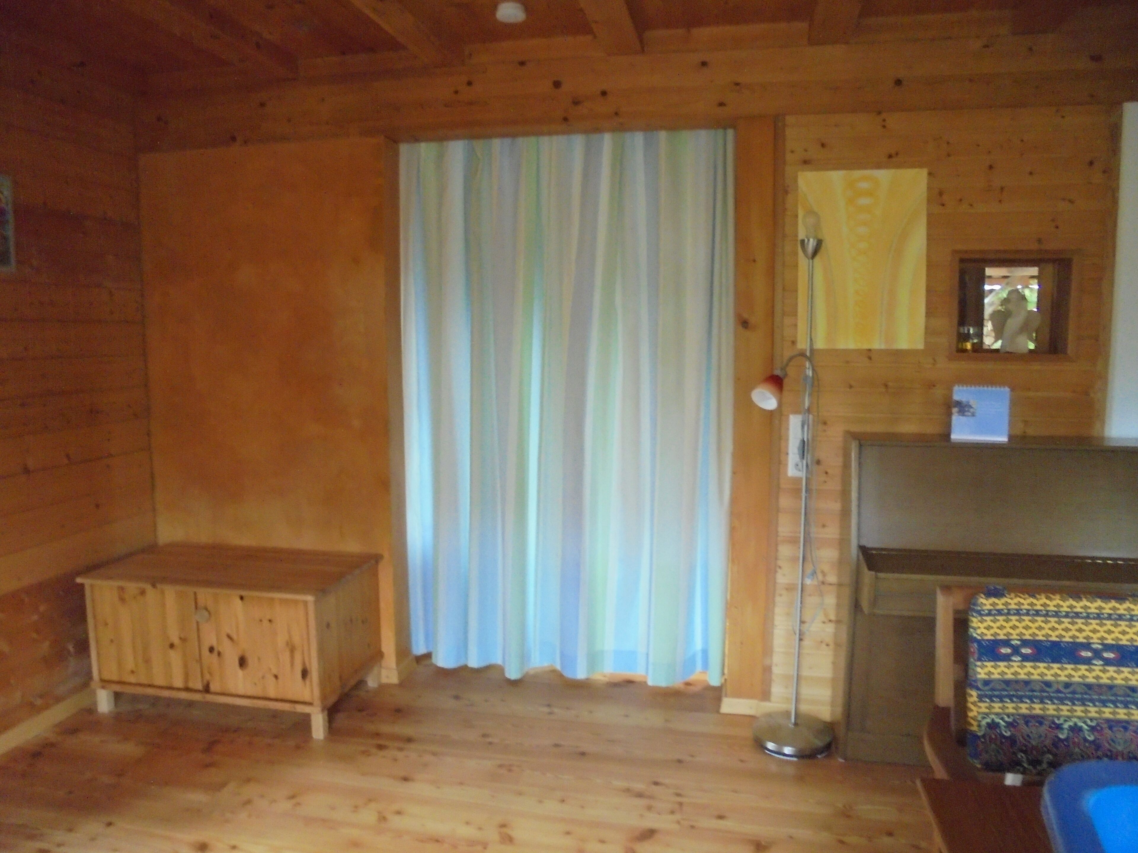 Interior de una cabaña de madera con un banco, una cortina, una lámpara de pie y una alfombra estampada.
