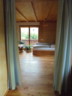 A wooden interior with a sofa and a dining table, a window with a view of greenery, and curtains on the sides. - Cozy apartment in a wooden house, perfect for hiking enthusiasts and active travelers (Ühlingen-Birkendorf)