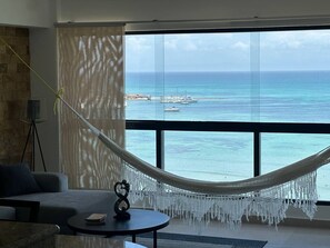 A room with a large window overlooking a beach and ocean. There is a hammock, a sofa, and a small table with a decorative item. - Beautiful 2-bedroom apartment with AC in Porlamar (Porlamar)