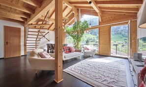 Ein geräumiges Zimmer mit Holzboden, einer Treppe, einem Sofa, einem Sessel und einem großen Fenster mit Blick auf eine grüne Landschaft.