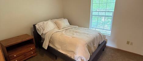 Un dormitorio con cama, mesita de noche, ventana con persianas y lámpara de techo.