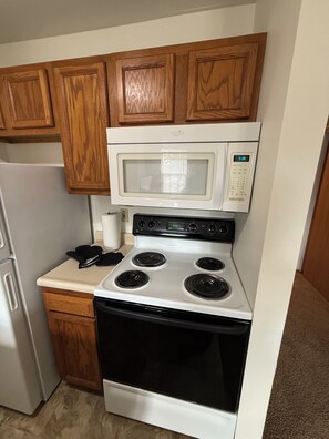 A compact kitchen with a white stove, oven, and microwave, wooden cabinets, and a white refrigerator. - Furnished 1BR Near OSU Reserved Parking + In-Unit Laundry (Columbus)