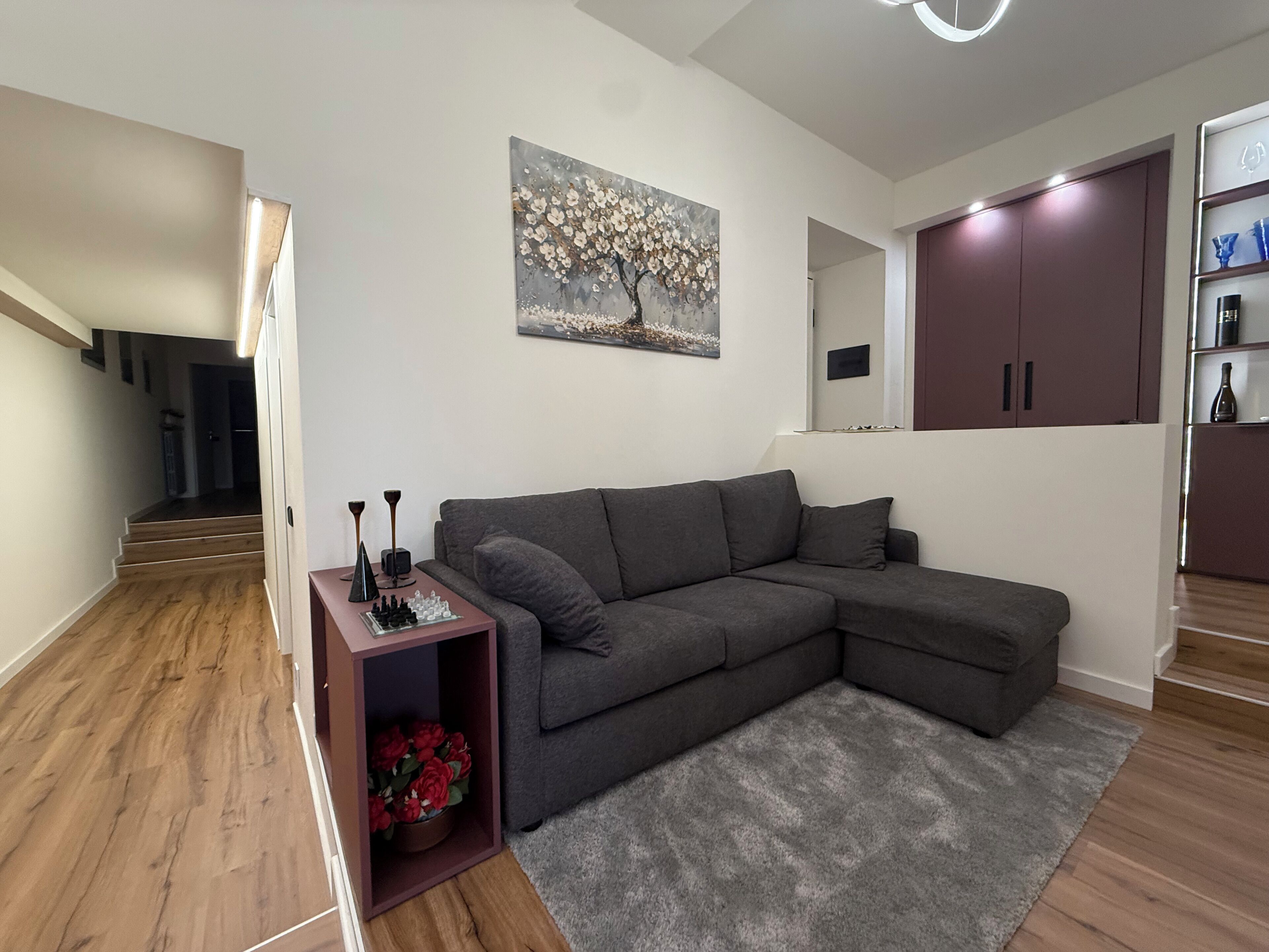 Una sala de estar moderna con un sofá seccional gris, una mesita con piezas de ajedrez, un cuadro de flores y una escalera que conduce a otra habitación.