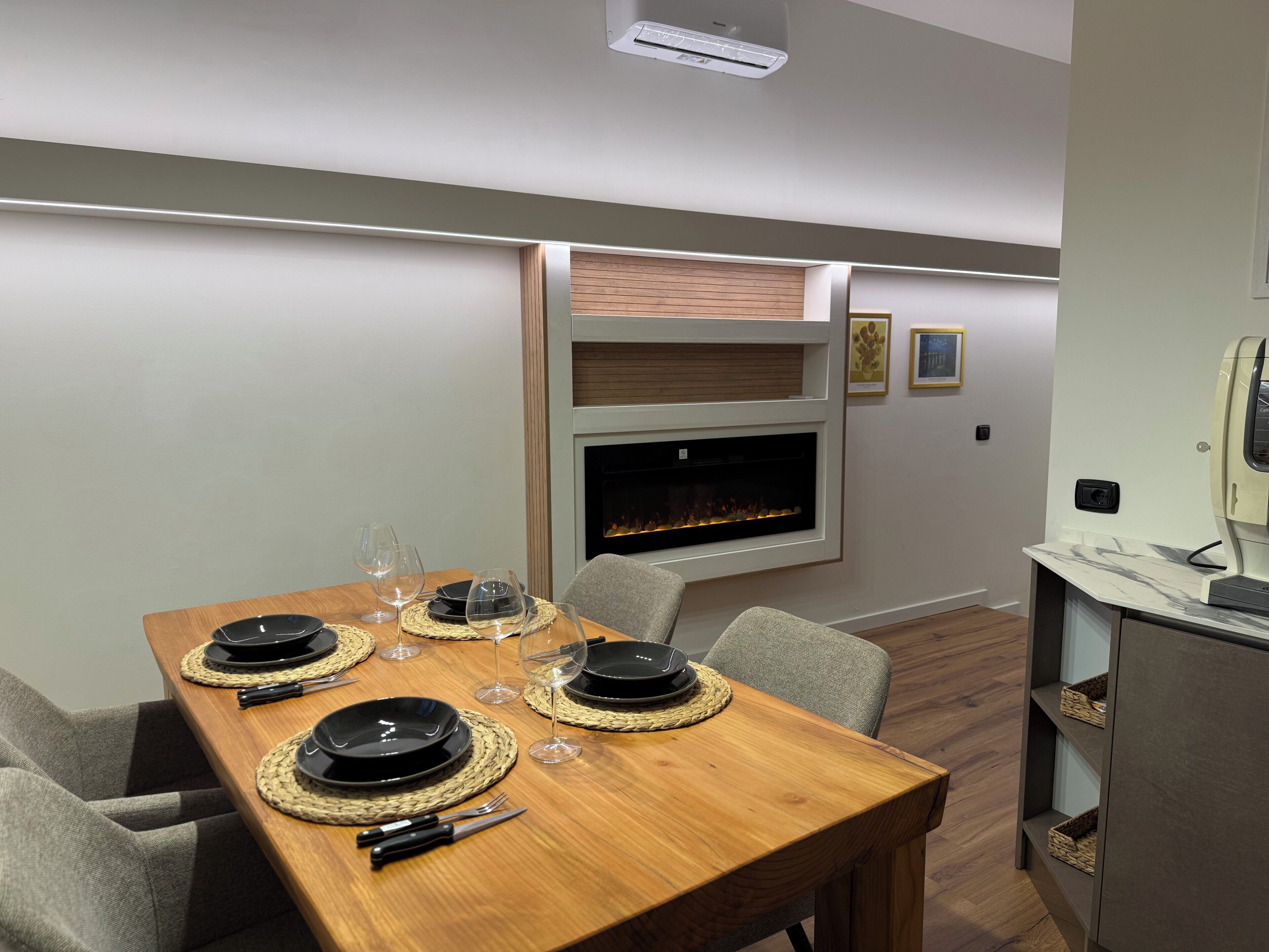 A dining area with a wooden table set for four, a fireplace, and a modern kitchen area.