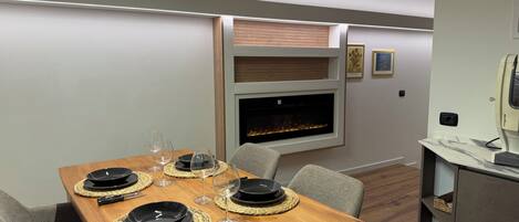 A dining area with a wooden table set for four, a fireplace, and a modern kitchen area.