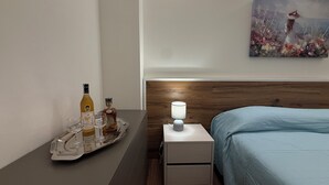 A hotel room with a bed, a nightstand with a lamp, a tray with bottles and glasses, and a painting on the wall.