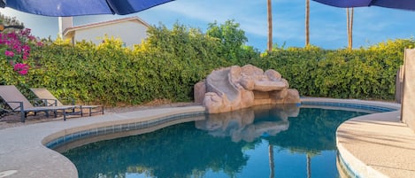 A swimming pool with a rock feature, surrounded by a hedge and umbrellas.