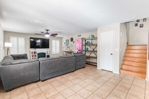 Ein Wohnzimmer mit grauem Ecksofa, einem Flachbildfernseher, einem Deckenventilator und einer Treppe, die zu einer weiteren Ebene führt.