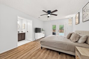 A bedroom with a bed, a ceiling fan, a TV, a desk, and a view of an outdoor area.