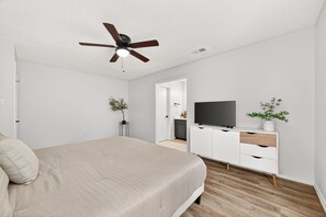 Une chambre à coucher moderne avec un lit, un ventilateur de plafond, un téléviseur posé sur un meuble blanc et une porte donnant accès à une salle de bain.