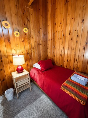 Ein Zimmer mit einer Holzwand, einem Bett mit roter Bettwäsche, einer roten Lampe auf einem Nachttisch aus Holz und einem kleinen weißen Eimer.