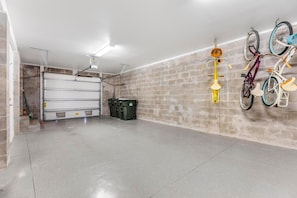 A garage with a concrete floor, a white garage door, and a brick wall. There are bicycles hanging on the wall and a green trash bin. - Supplication at Lost Key Golf & Beach Club (Pensacola)