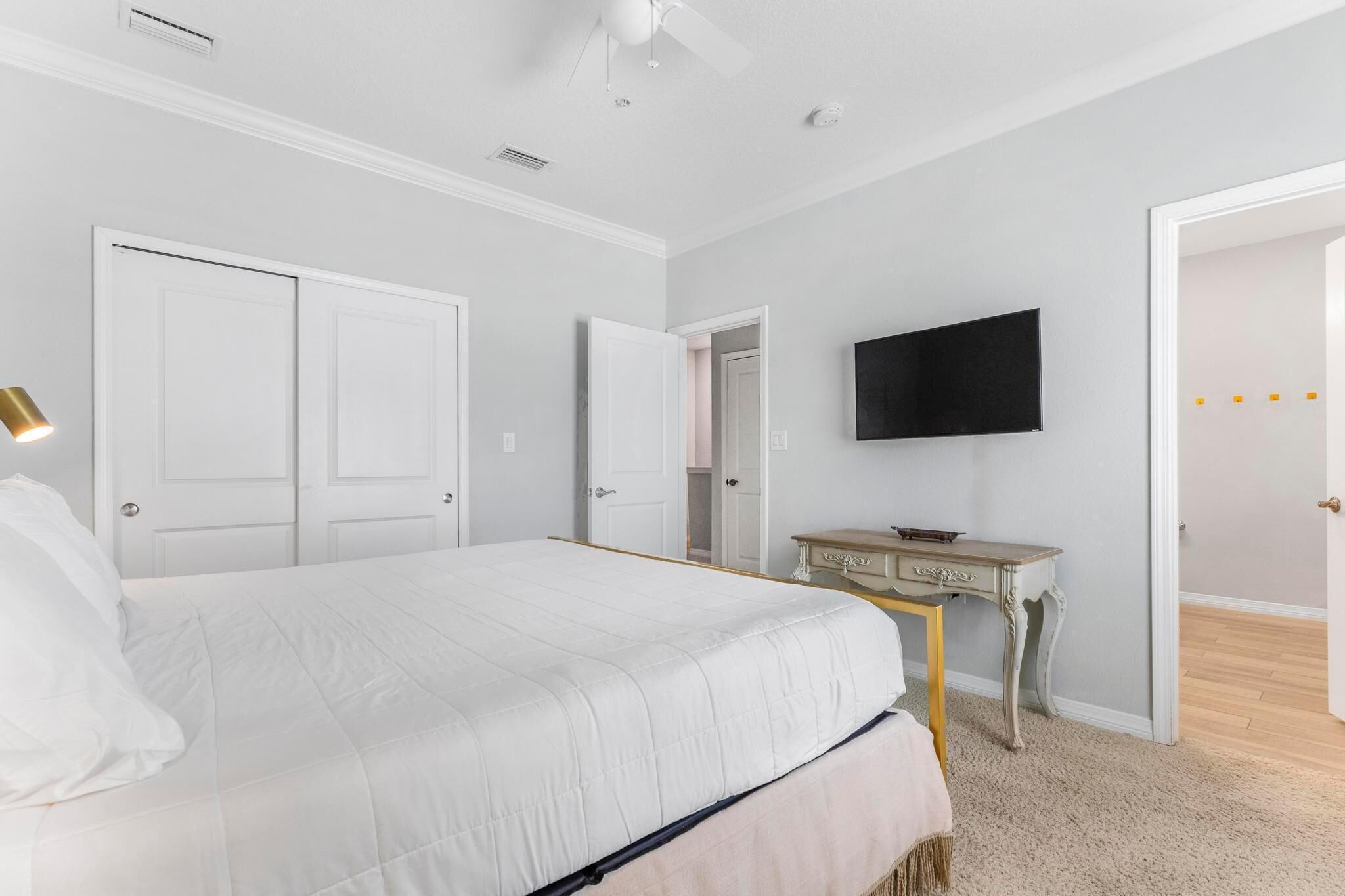A bedroom with a bed, a desk, a television, and a door leading to another room.