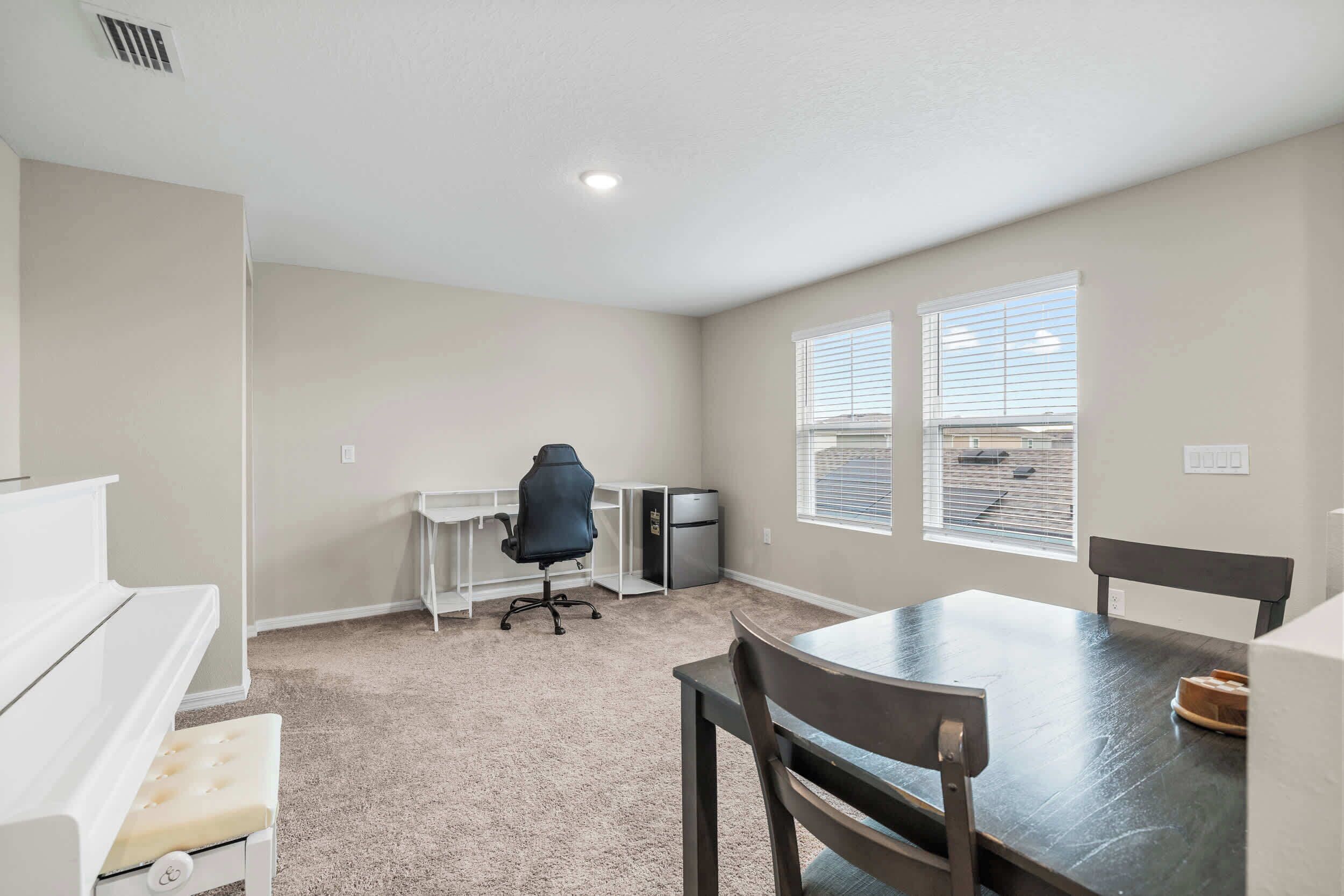 A small, well-lit room with a kitchenette, a single office chair, a dining table with chairs, and a window with blinds.