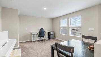 A small, well-lit room with a kitchenette, a single office chair, a dining table with chairs, and a window with blinds.