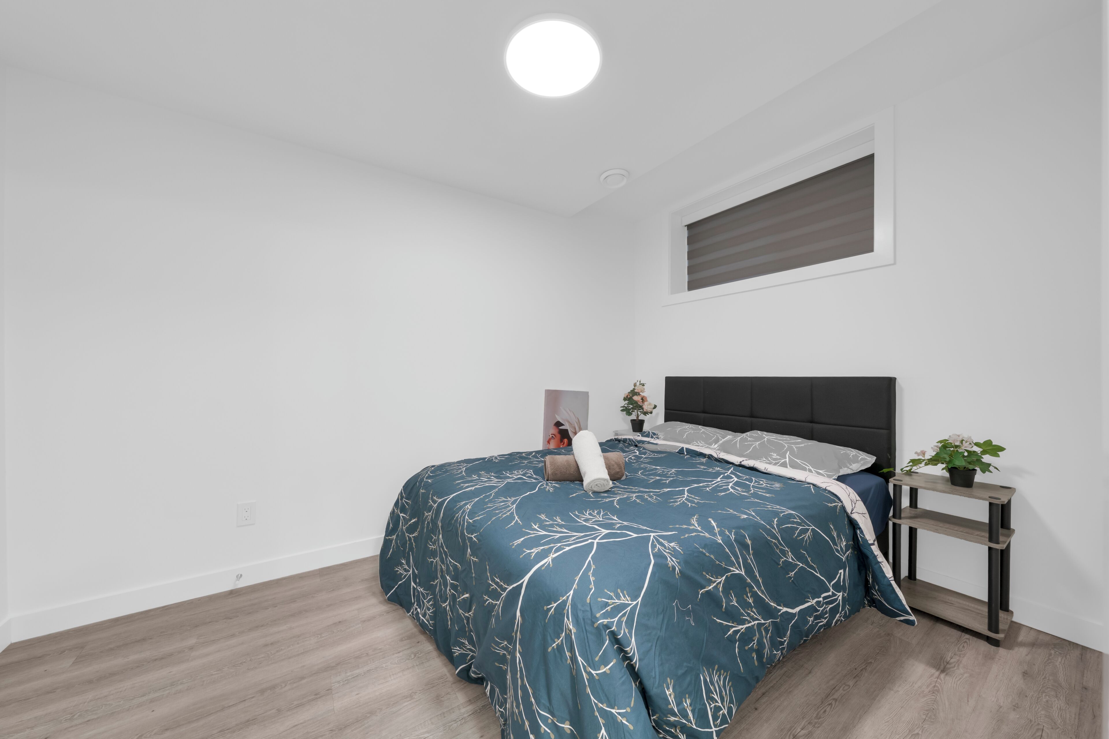 A bedroom with a bed, a bedside table, a potted plant, and a window with blinds.