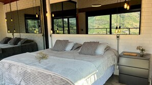 A bedroom with a large bed, a nightstand, and a window with a view of greenery. - Vista Baú: Luxury Chalet with Breathtaking Views of Pedra do Baú (São Bento do Sapucaí)