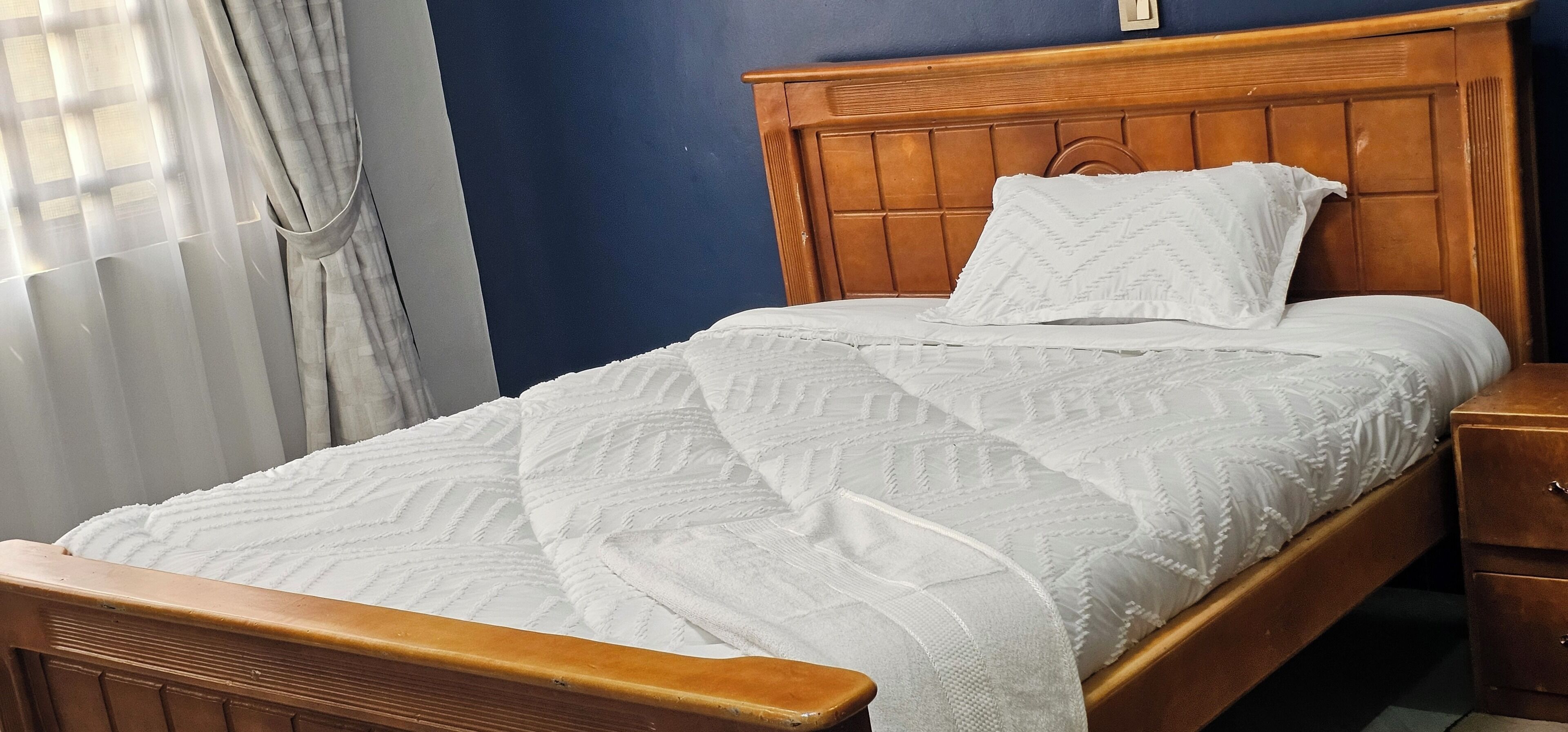 Ein Einzelbett mit Holz-Kopf- und Fußteil, einer weißen Steppdecke und einem Nachttisch mit Schublade.