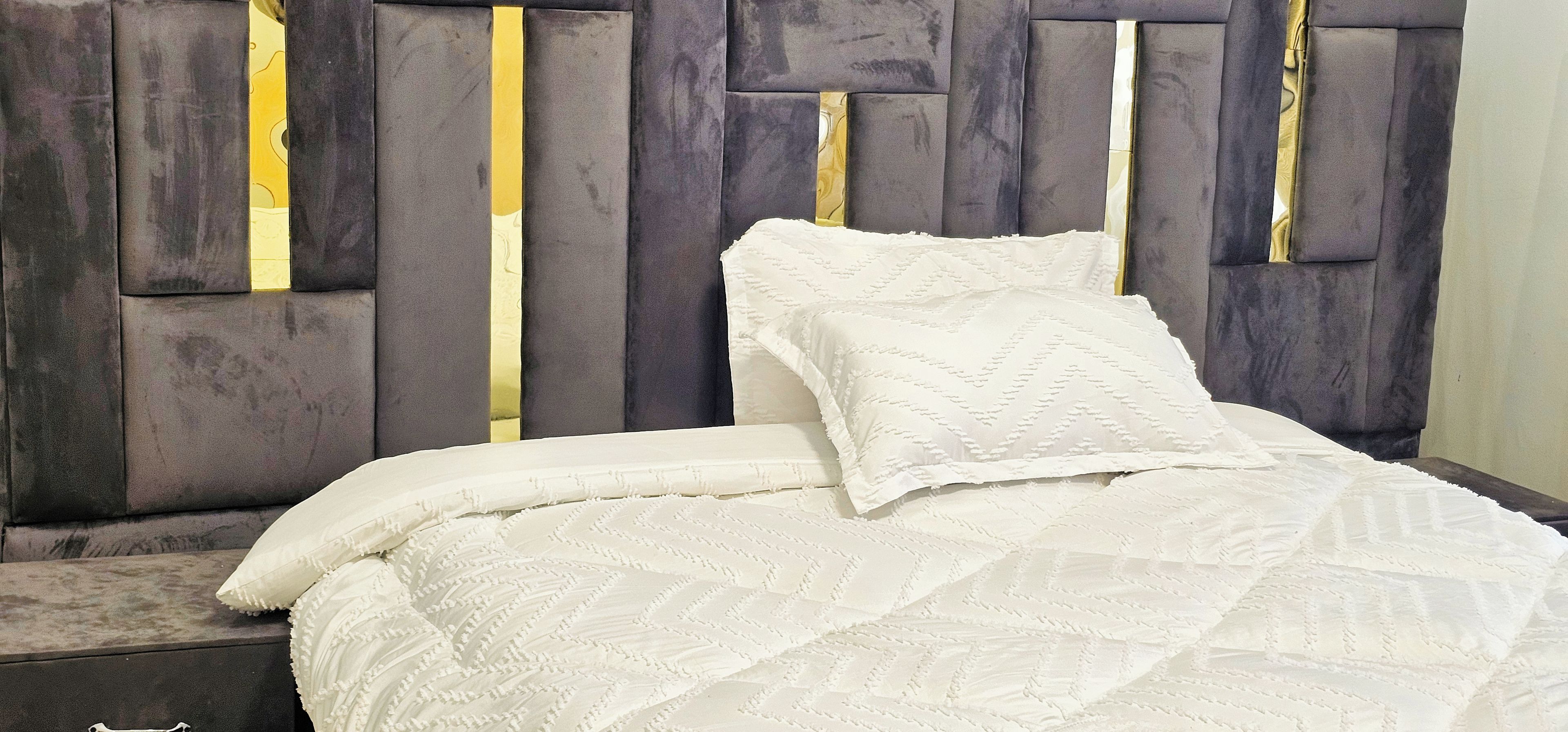 Una cama con una colcha blanca texturizada y almohadas, un cabecero con un diseño geométrico y una mesita de noche con una lámpara.