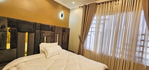 A hotel room with a large bed, a headboard with light fixtures, and a window with curtains. - Babz Residence, A private countryside retreat in a serene natural setting (Lutaba Trading Centre)