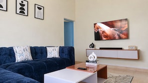 A living room with a dark blue sofa, a coffee table, a flat-screen TV mounted on the wall, and a bookshelf with decorative items.