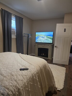 A bedroom with a bed, a television on a stand, a fireplace, a door labeled 'B', and a window with curtains.