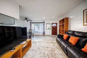 A living room with a television, a wooden coffee table, a leather sofa with orange cushions, a ceiling fan, and a view of the outside through a window. - Pretty apartment (Los Angeles)