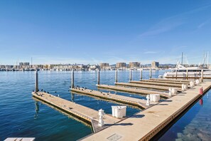 Un port esportiu amb vaixells amarrats, un moll amb un far, i un skyline urbà al fons.