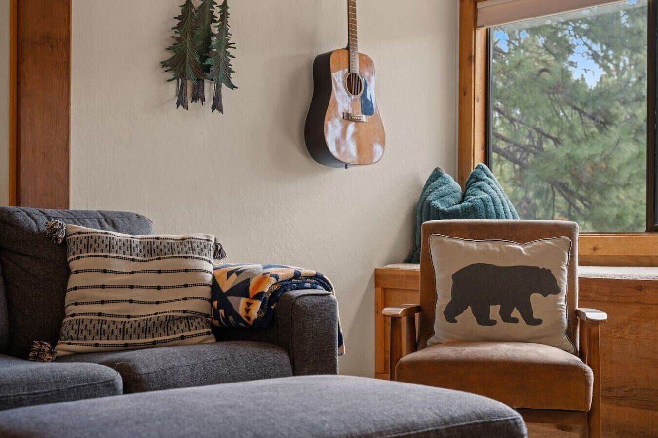 Una sala de estar acogedora con un sofá gris, un sillón de madera con un cojín de oso, una guitarra en la pared y una ventana con vistas a árboles.