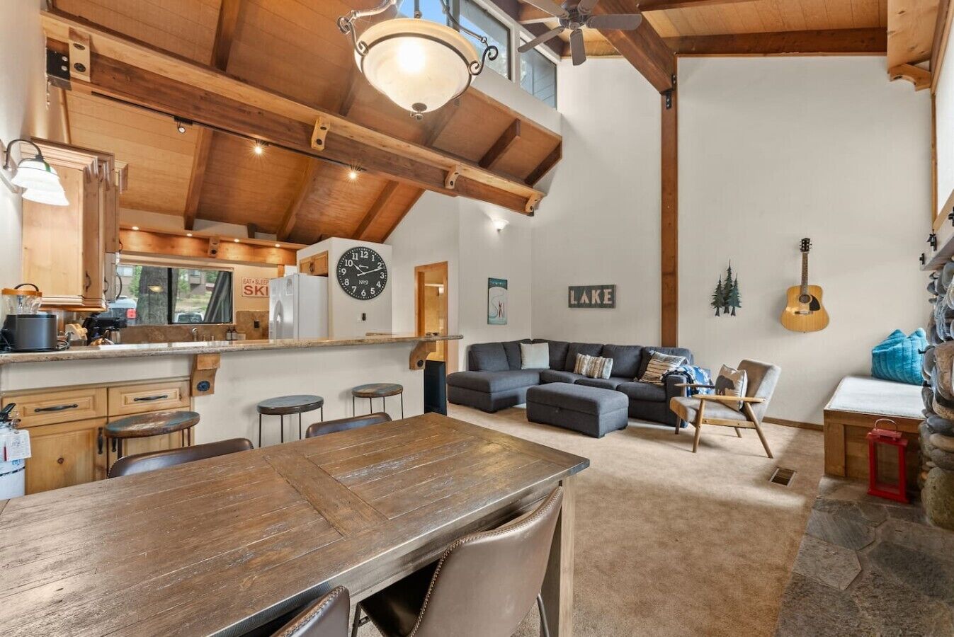 Un vaste espace de vie comprenant une grande table à manger en bois, un ensemble de canapés et une cuisine avec des poutres apparentes et une horloge.