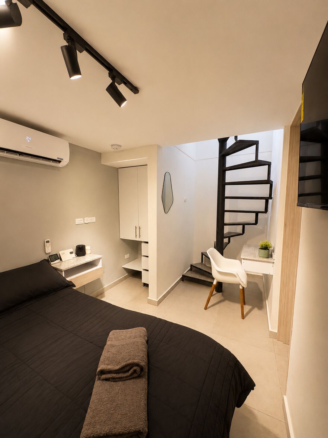 A modern bedroom with a bed, bedside table, and a desk with a chair. There is a staircase with a unique design.