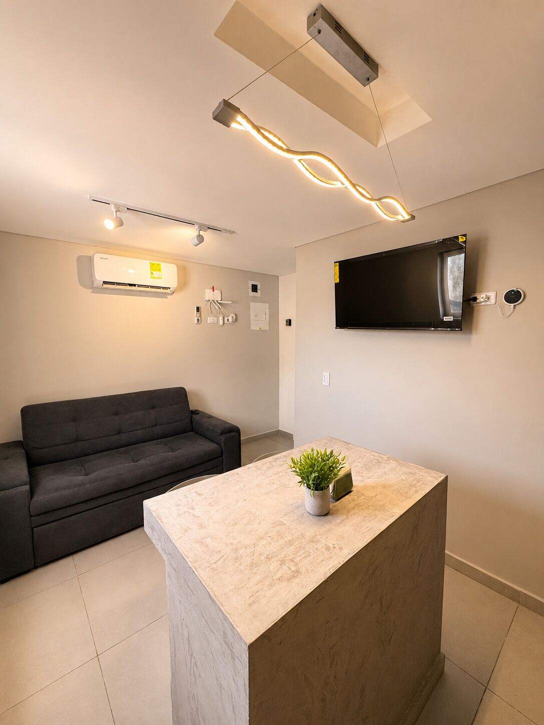 Un salon moderne avec un canapé, un téléviseur à écran plat fixé au mur, un luminaire suspendu au plafond et une petite plante en pot posée sur une table en marbre.