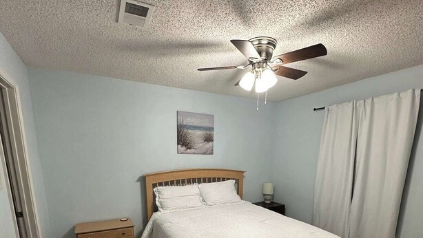 Une chambre à coucher avec un lit, une table de chevet, un ventilateur de plafond et un tableau accroché au mur.