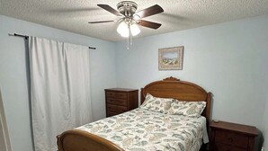 Une chambre à coucher avec un lit en bois, un ventilateur de plafond, une table de chevet, une commode et une fenêtre avec des rideaux.