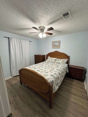 Een slaapkamer met een houten bed, een plafondventilator, een nachtkastje, een dressoir en een raam met gordijnen.