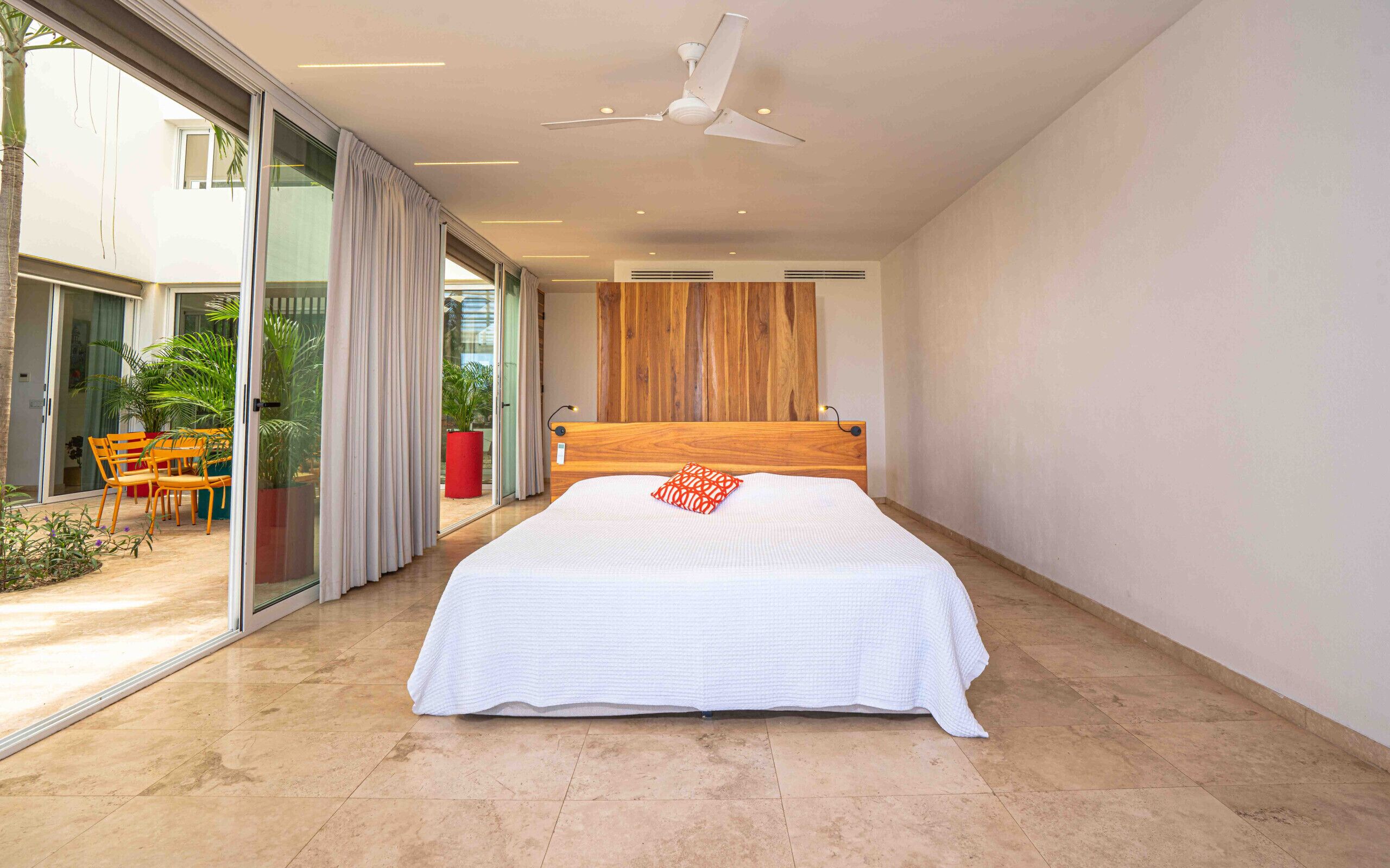 A modern bedroom with a large bed, a ceiling fan, and a sliding glass door leading to an outdoor area with plants and furniture.