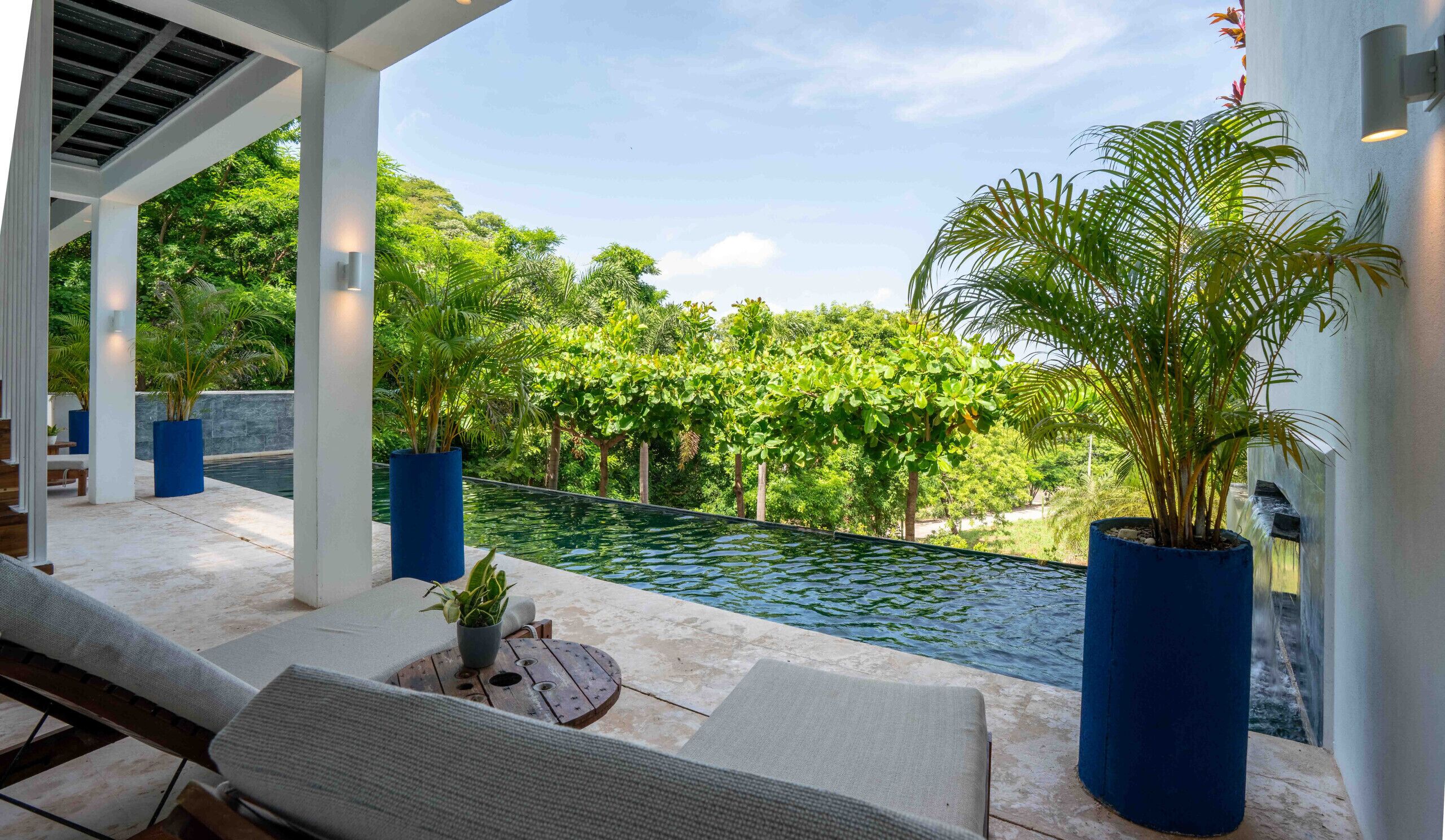 A poolside area with a lounge chair, a small table with a plant, and a view of a swimming pool surrounded by lush greenery.