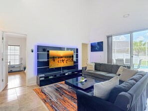 A modern living room with a flat-screen TV mounted on a wall, a sofa, a coffee table, and a view of the outdoors through large windows. - Marvelous 7-bedroom house with AC in charming Kissimmee (Kissimmee)