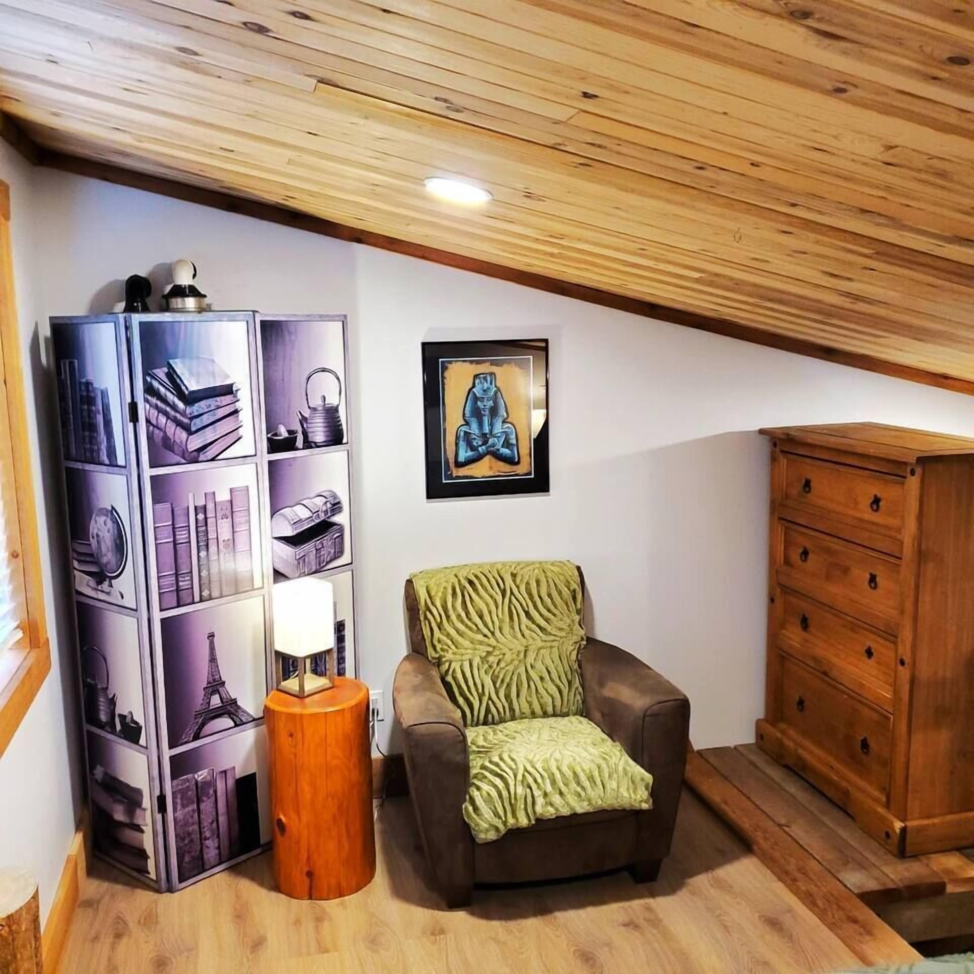 A cozy room with a wooden ceiling, a bookshelf with travel-themed books, a green chair, a wooden cabinet, and a framed picture on the wall.