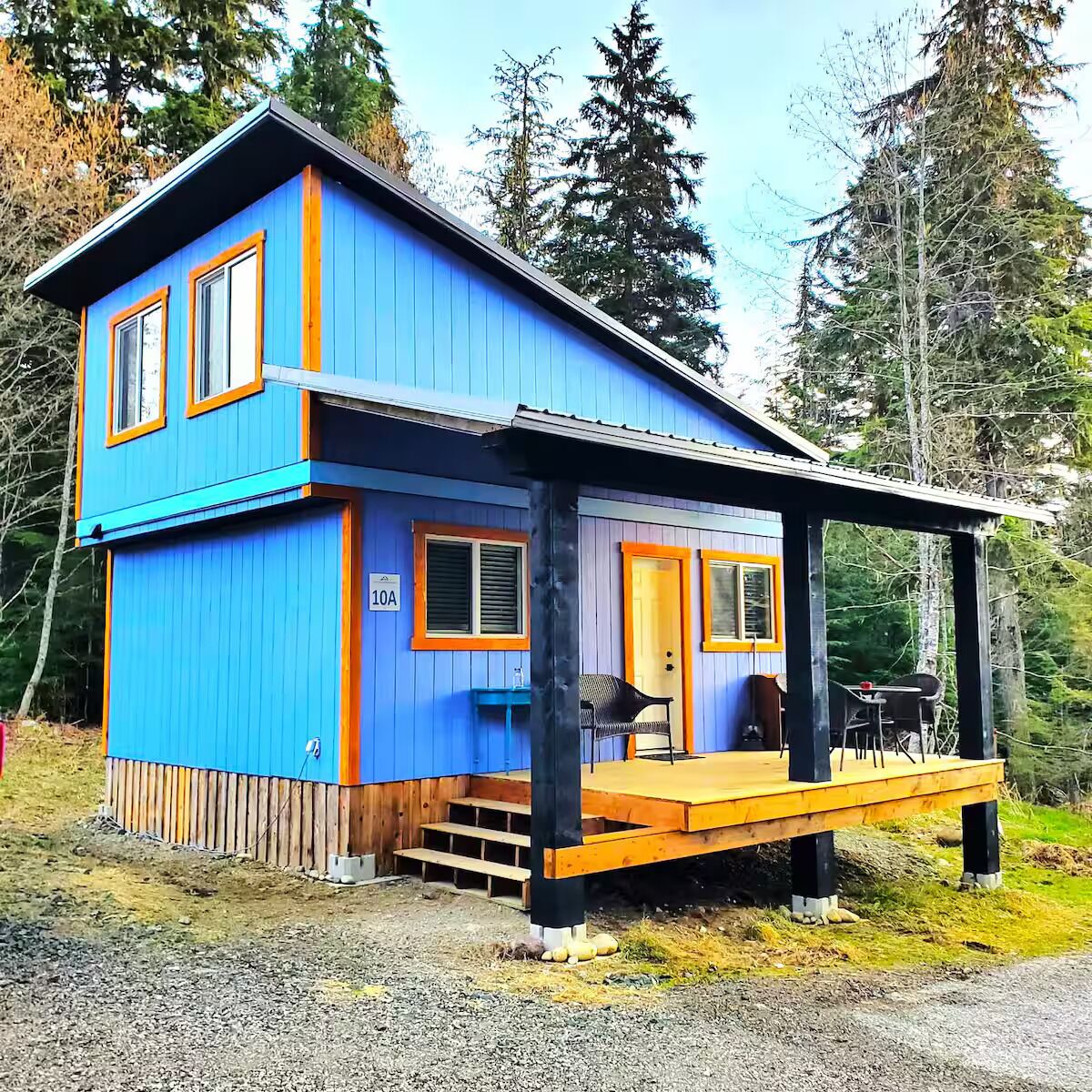 Una cabaña azul de dos pisos con terraza de madera, un porche y un pequeño conjunto de mesa y sillas.