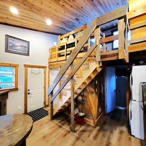 A rustic cabin interior with a wooden staircase leading to a loft, a kitchen area with a refrigerator, and a dining table.