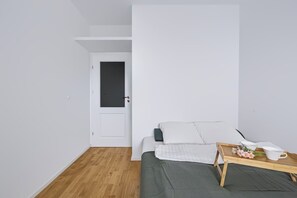 Una habitación pequeña y minimalista con una cama, una mesita de noche con lámpara y una puerta que da a otra estancia.