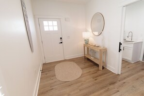 Um corredor com piso de madeira, um tapete redondo, um console com abajur, um espelho e uma porta com janela.