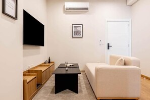 Una sala de estar moderna con un televisor de pantalla plana, un cuadro enmarcado, una mesa de centro negra, un sofá beige y una puerta blanca.