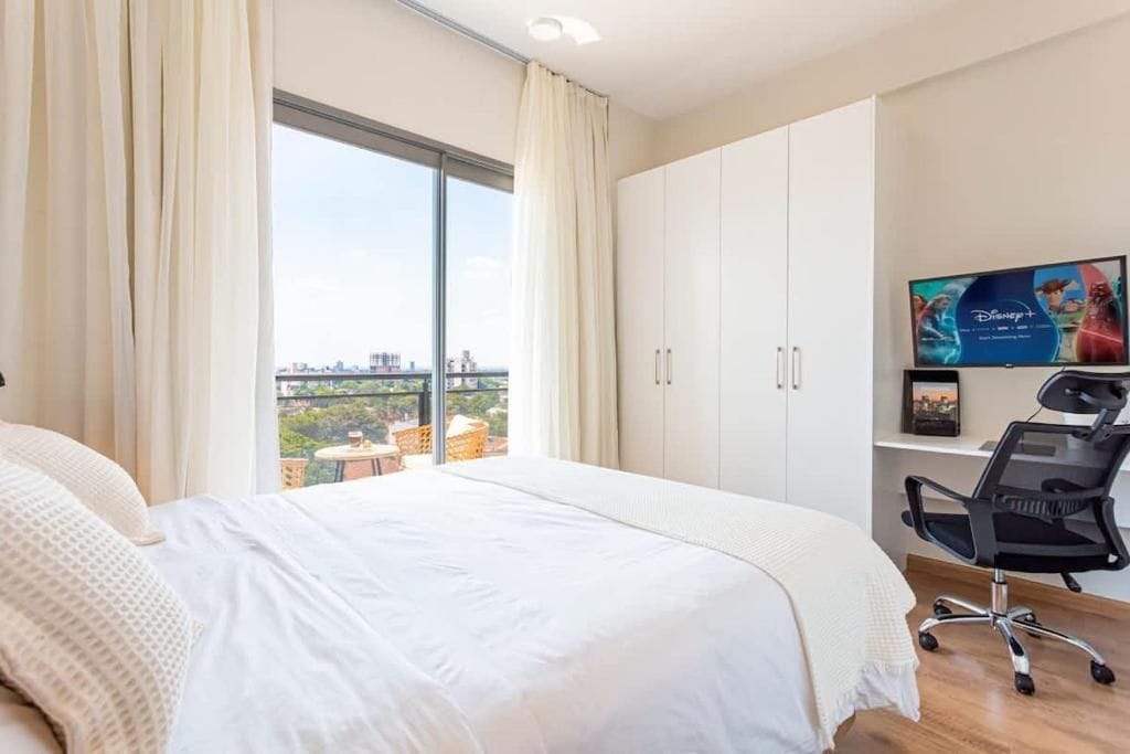 A hotel room with a large bed, a desk with a chair, a television, and a view of the city through a large window.